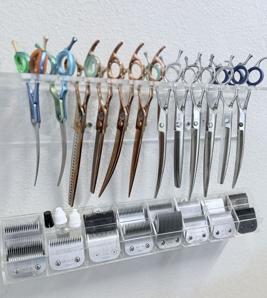 A wall-mounted acrylic rack holding about a dozen grooming shears, above a tray of clipper blades in numbered sizes.