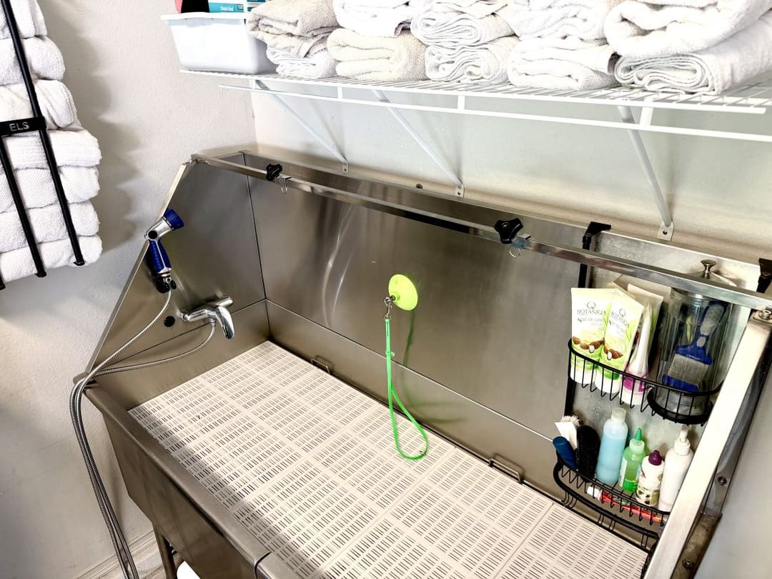 A stainless steel bathing tub with a sprayer, a grooming loop and caddies of shampoo and brushes, under a wire shelf of folded white towels.