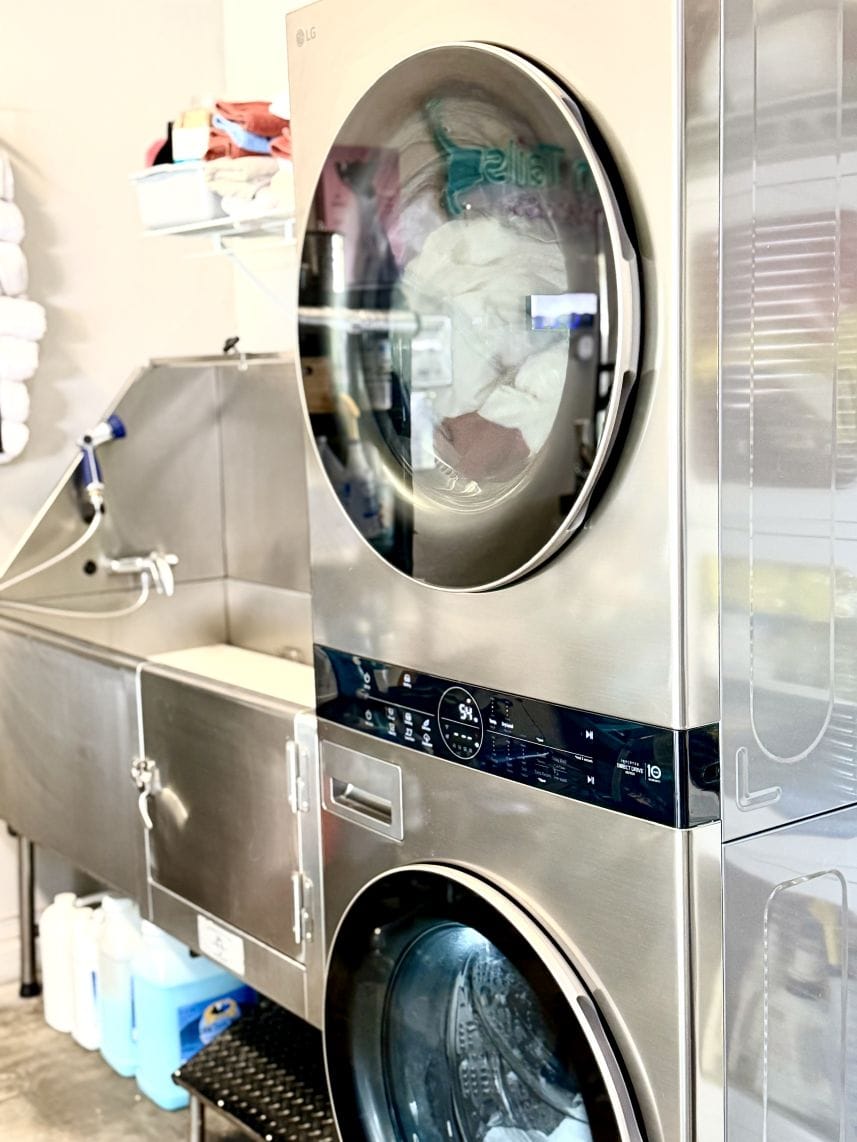 A stacked washer and dryer beside the bathing tub, towels visible through the dryer door, and jugs on a shelf underneath the tub.