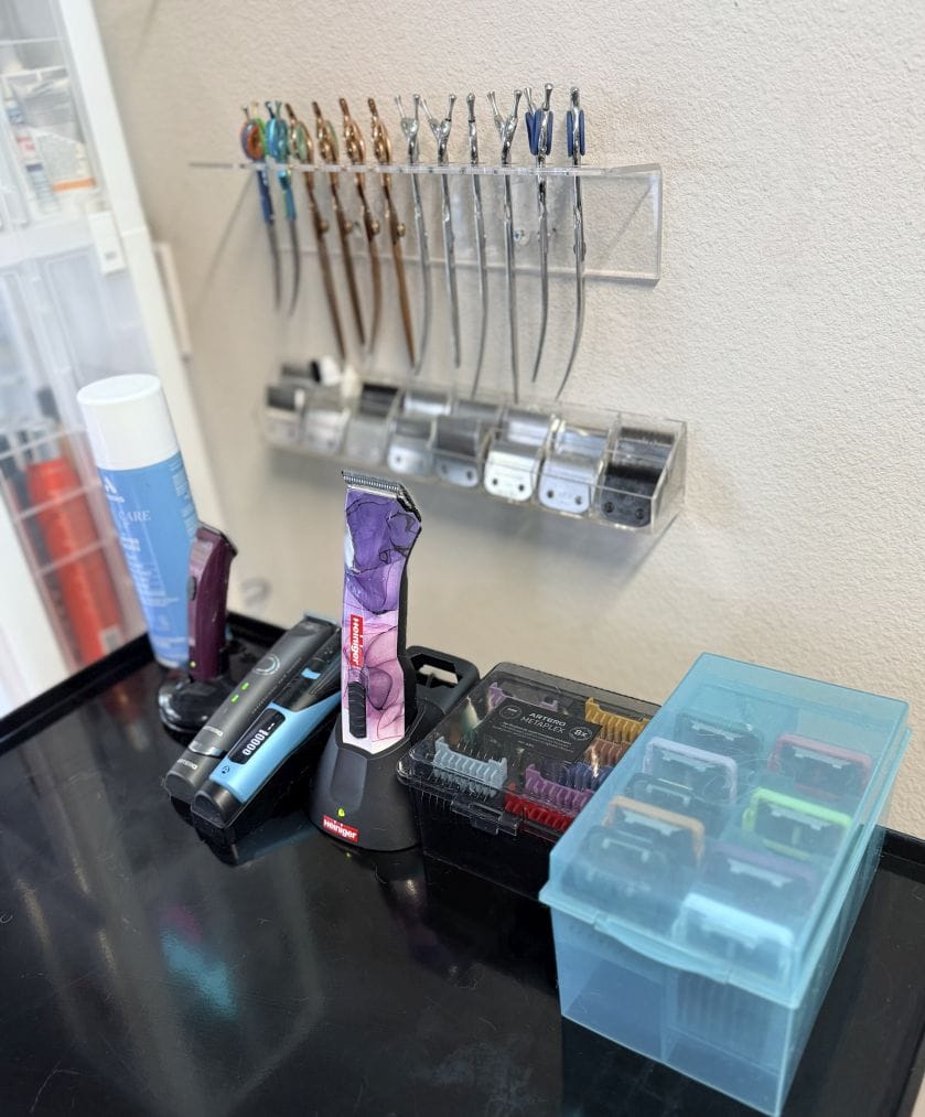 Cordless clippers standing on a charging base on a black counter, a case of guard combs beside them, and more shears and blades on the wall above.