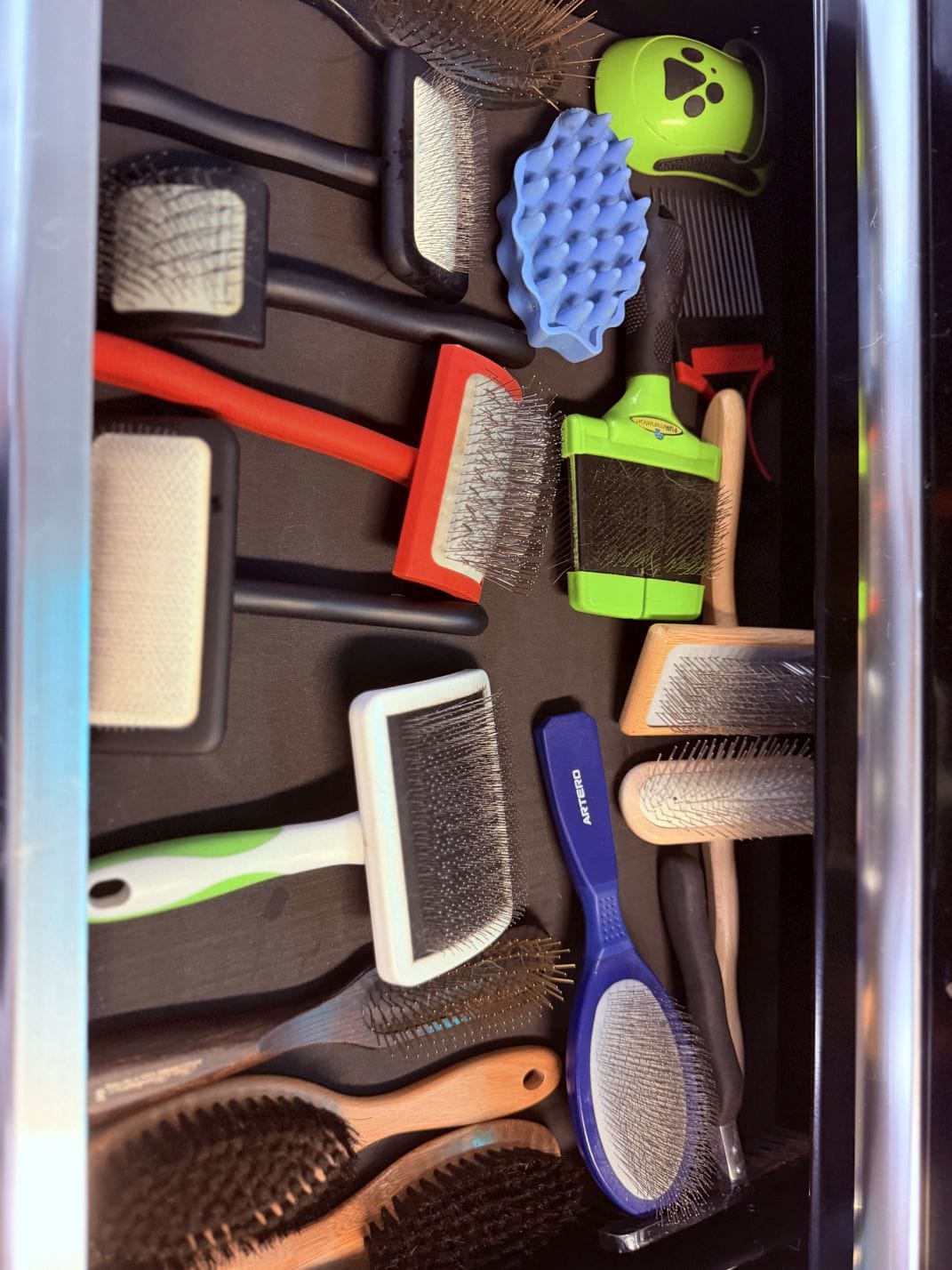 An open drawer of grooming brushes and combs, slicker brushes, pin brushes, a rubber curry and a metal comb among them.